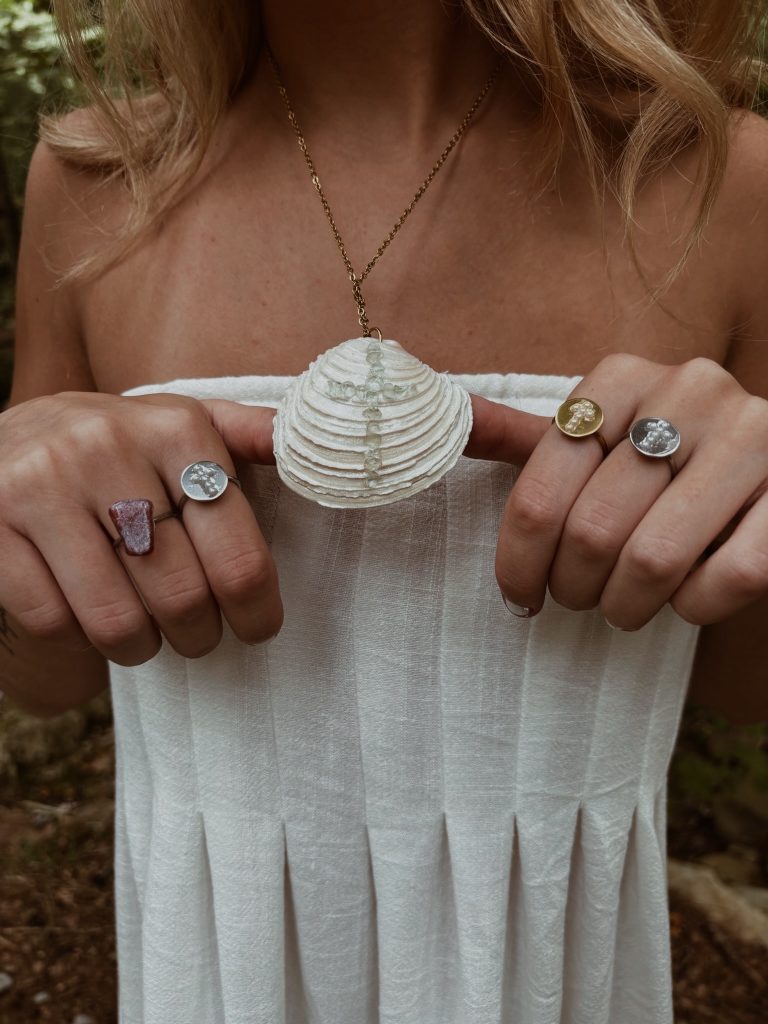 A white scallop shell pendant on a gold chain, held up between two hands wearing pearl and raw-stone rings