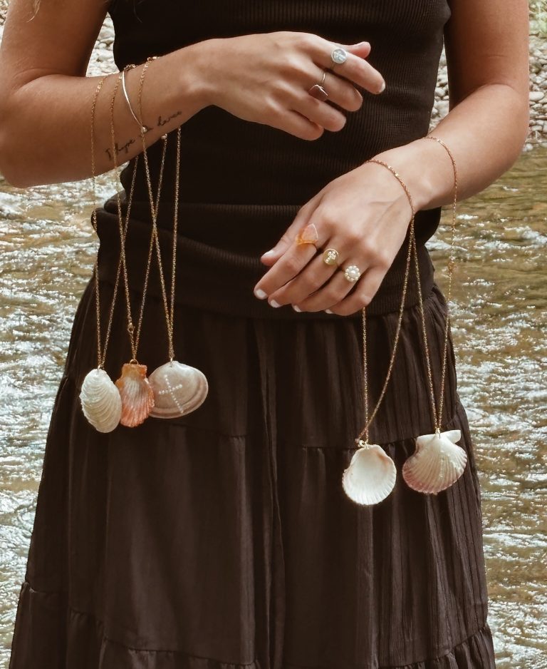 Five shell pendants and fine gold chain bracelets worn together beside a creek
